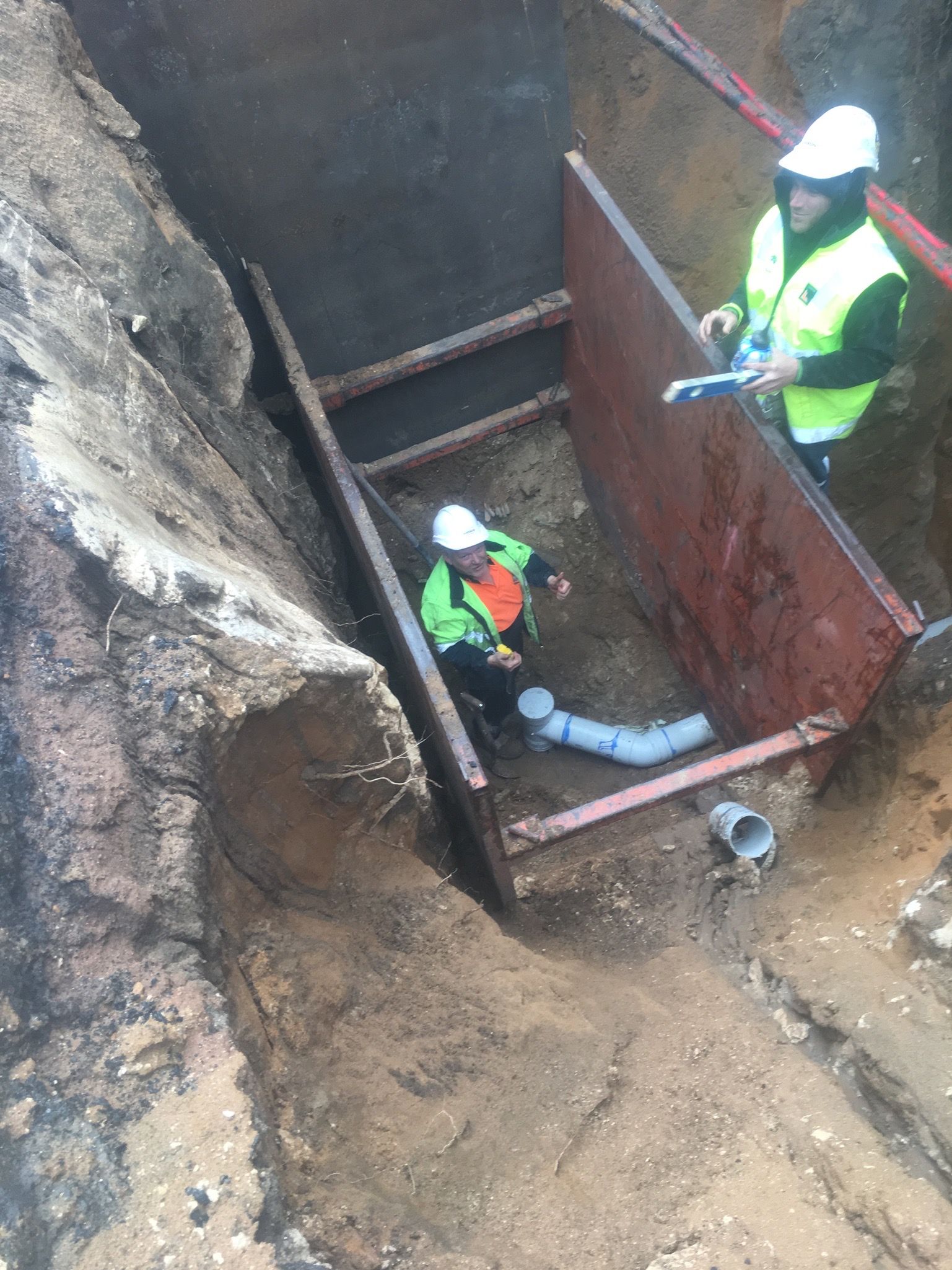 Two workers in hi-vis installing pipework inside a shored excavation