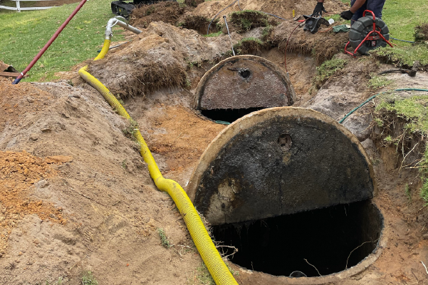 An old round tank uncovered in sandy soil with a yellow suction hose beside it